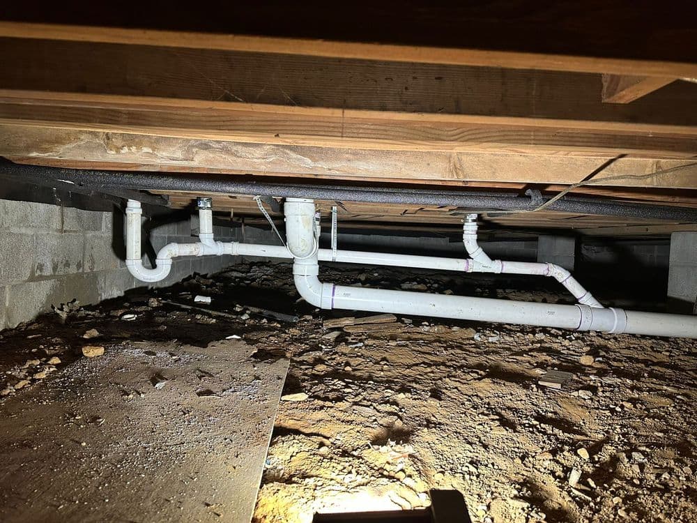 Visible plumbing in a dimly lit crawl space with dirt and debris on the ground.