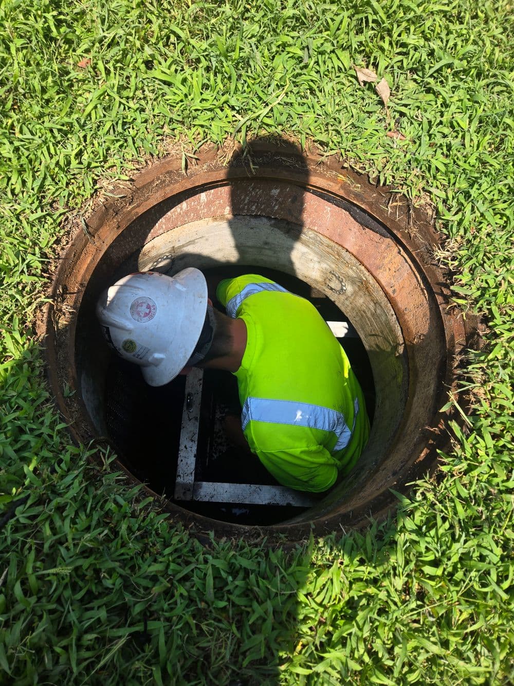 Worker in safety gear inspecting a manhole in a grassy area on a sunny day.