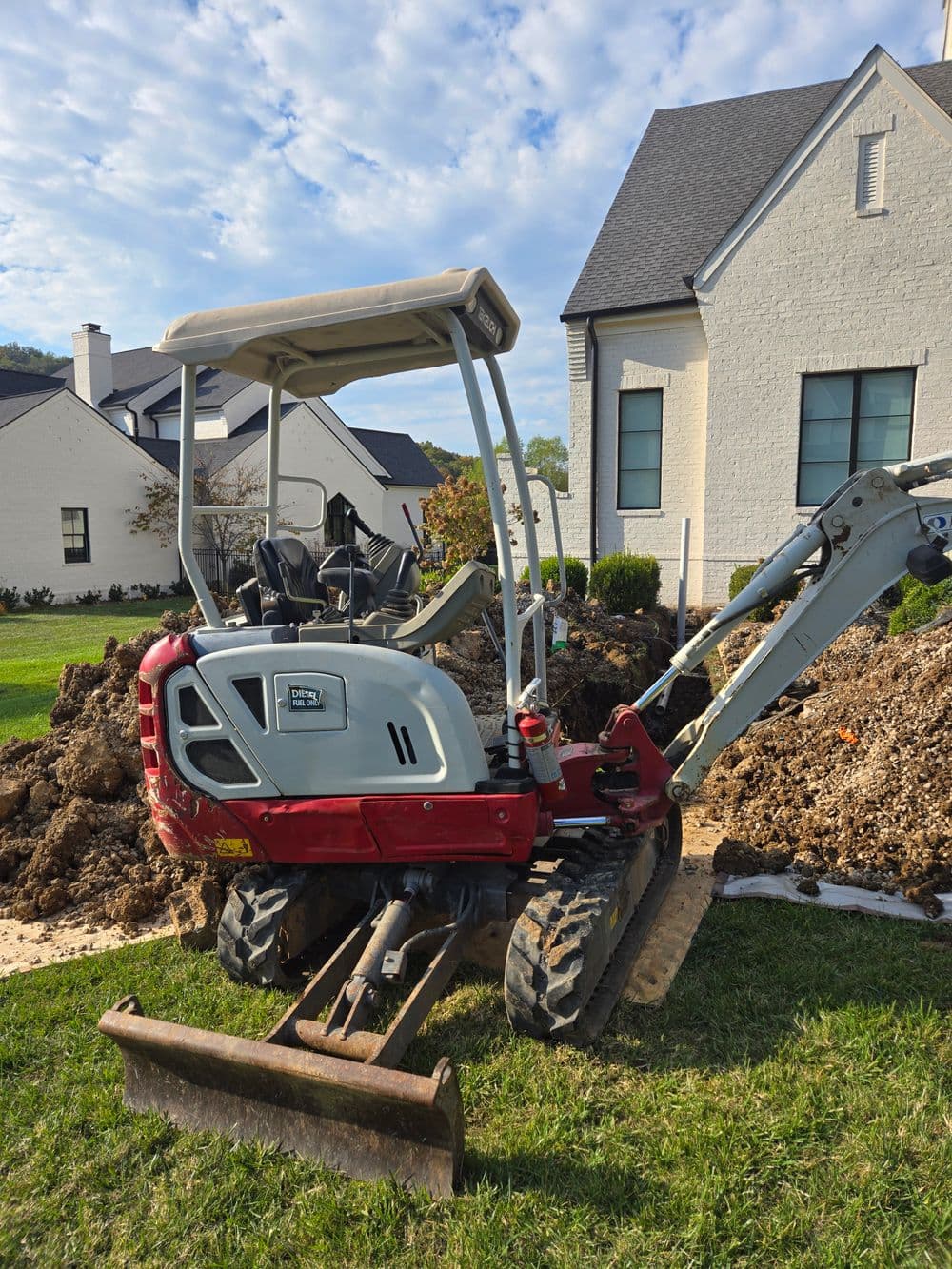 Small excavator on a construction site near a modern home, surrounded by dug earth and greenery.