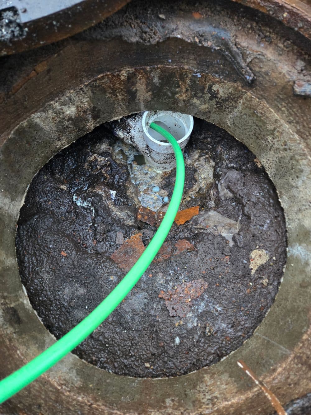 Close-up of a drainage pipe surrounded by murky water and debris, topped with a green hose.