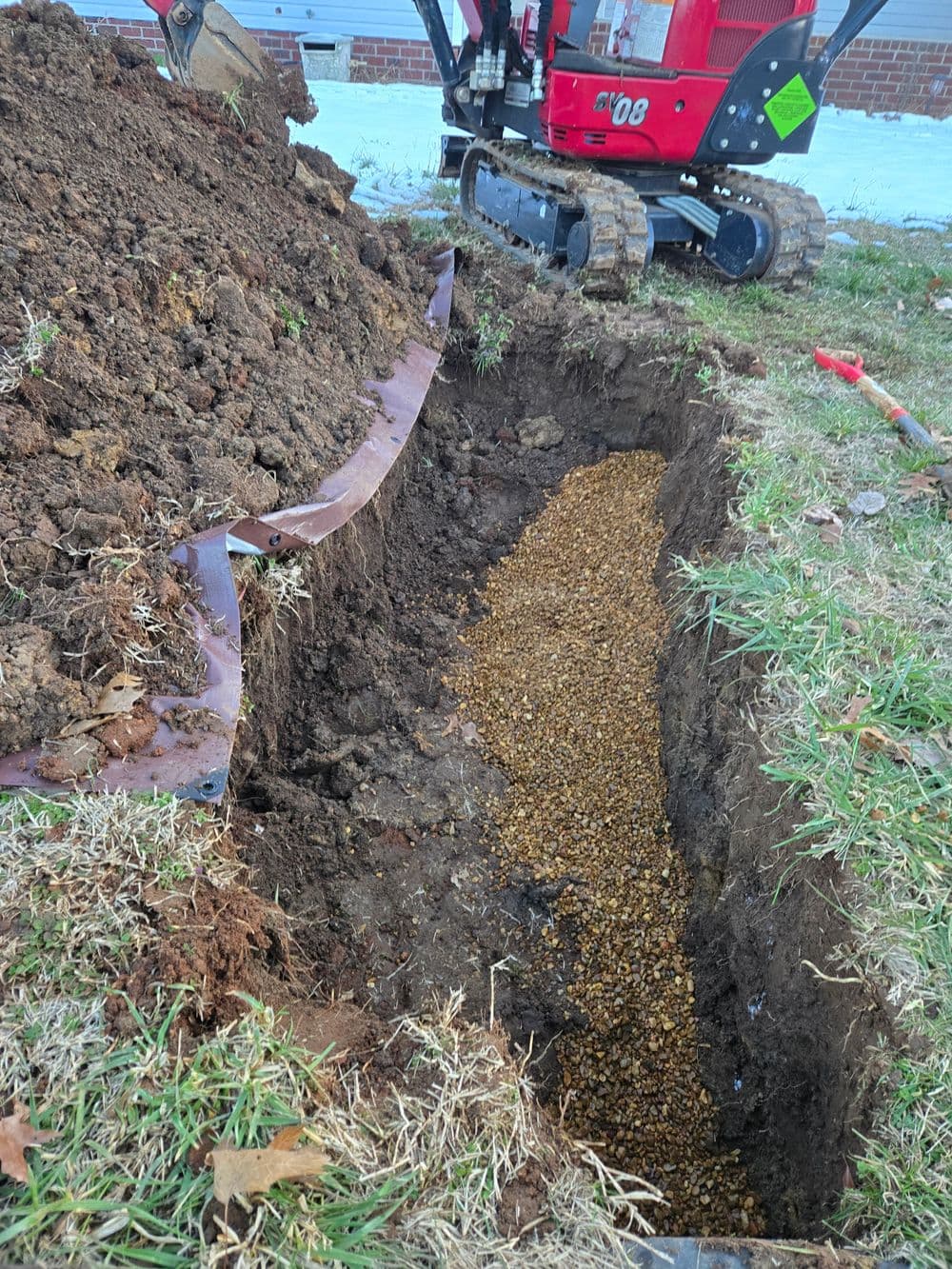 Excavated trench filled with gravel near a mini excavator in a yard.