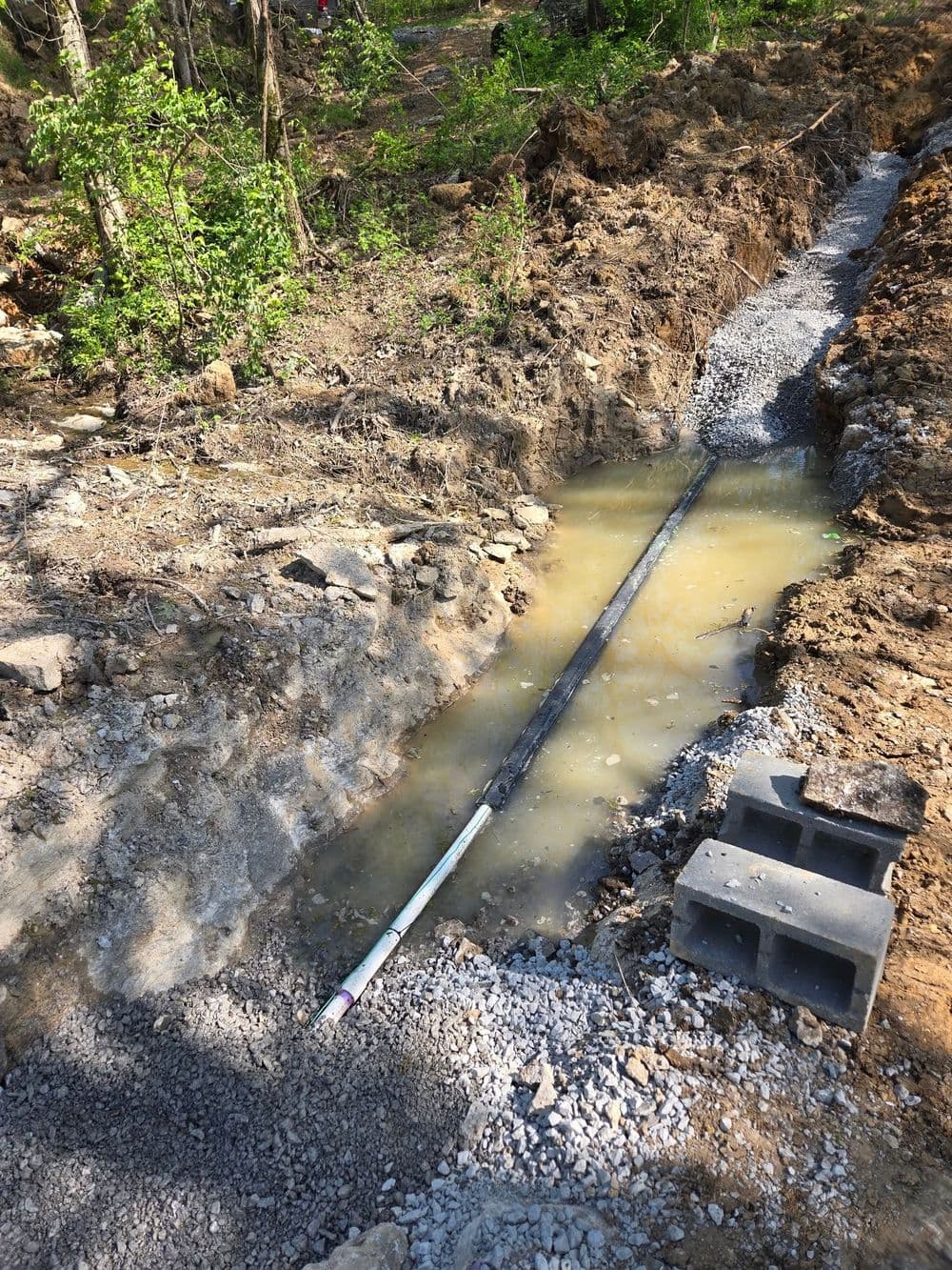 Excavated area with water, gravel, and a pipeline under construction in a wooded location.