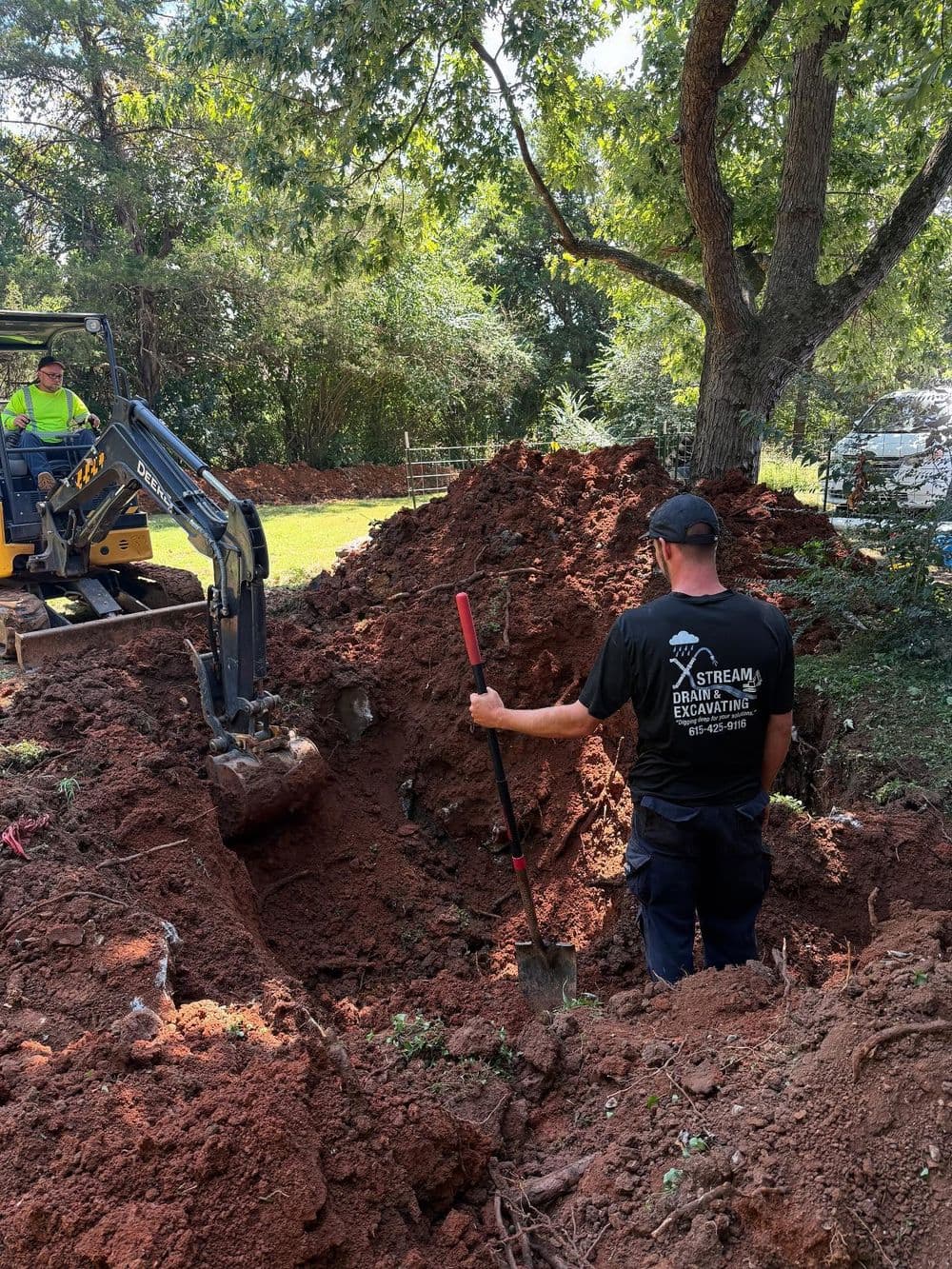 Excavation work in progress with a backhoe and worker digging a large trench in soil.