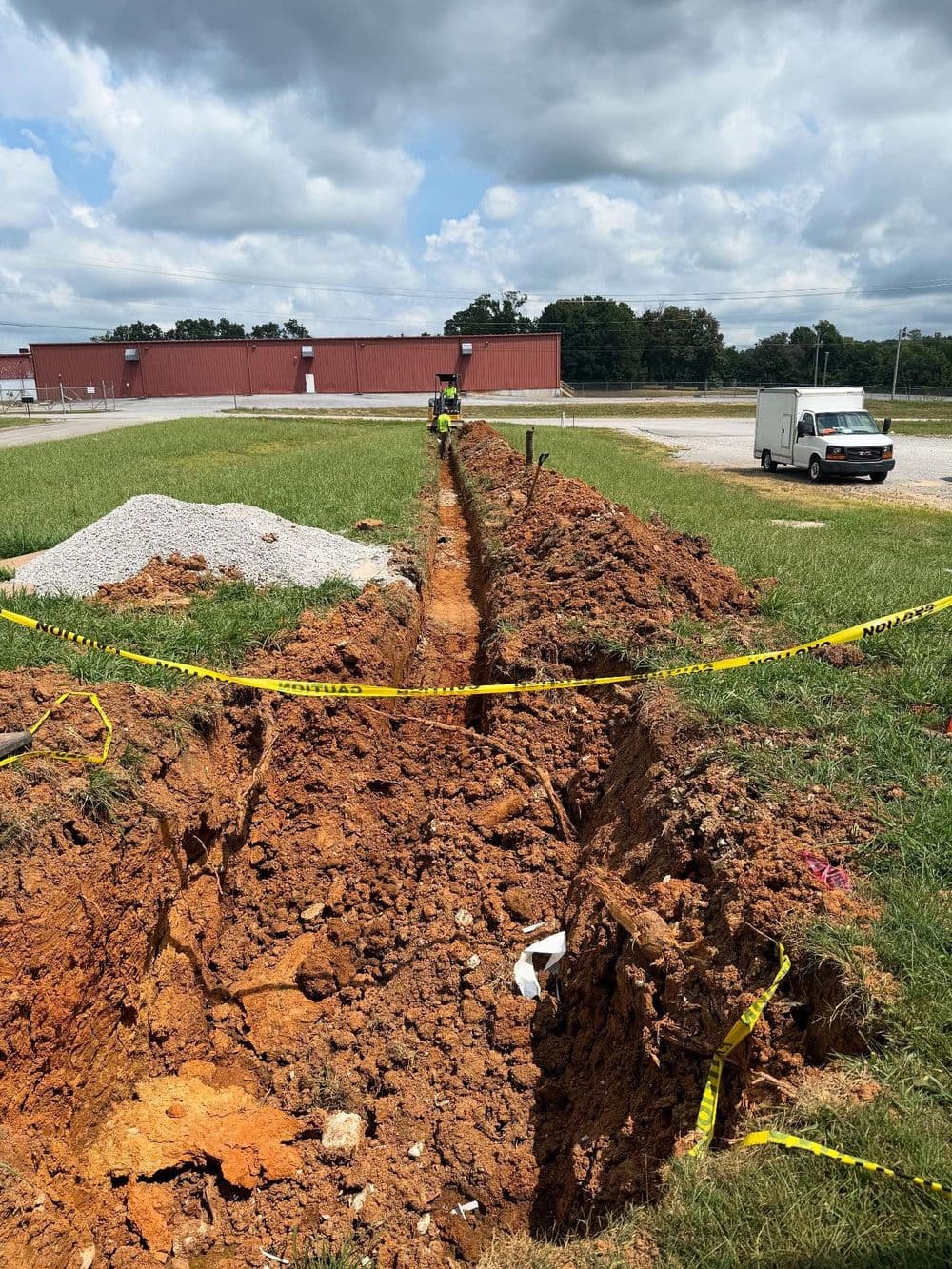 Construction workers digging a trench in a grassy area with safety barriers and equipment nearby.