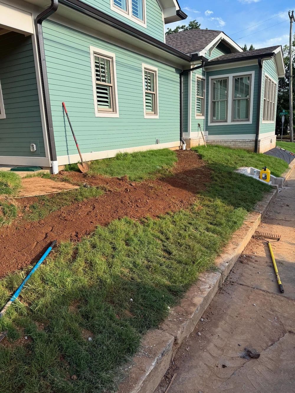 Landscaping project with freshly turned soil and tools beside a green house foundation.