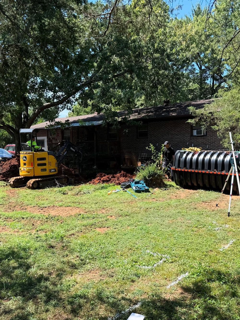 Excavator digging near a home, with construction materials and a green lawn visible.