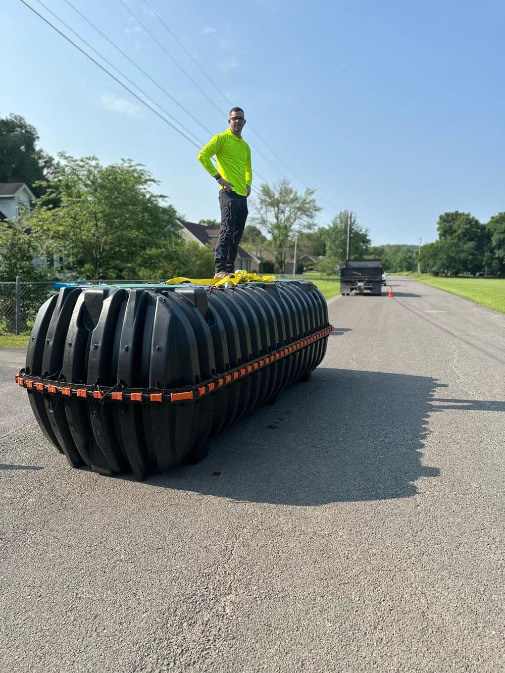 Worker standing atop large black plastic tank on a road, surrounded by trees and sunny sky.