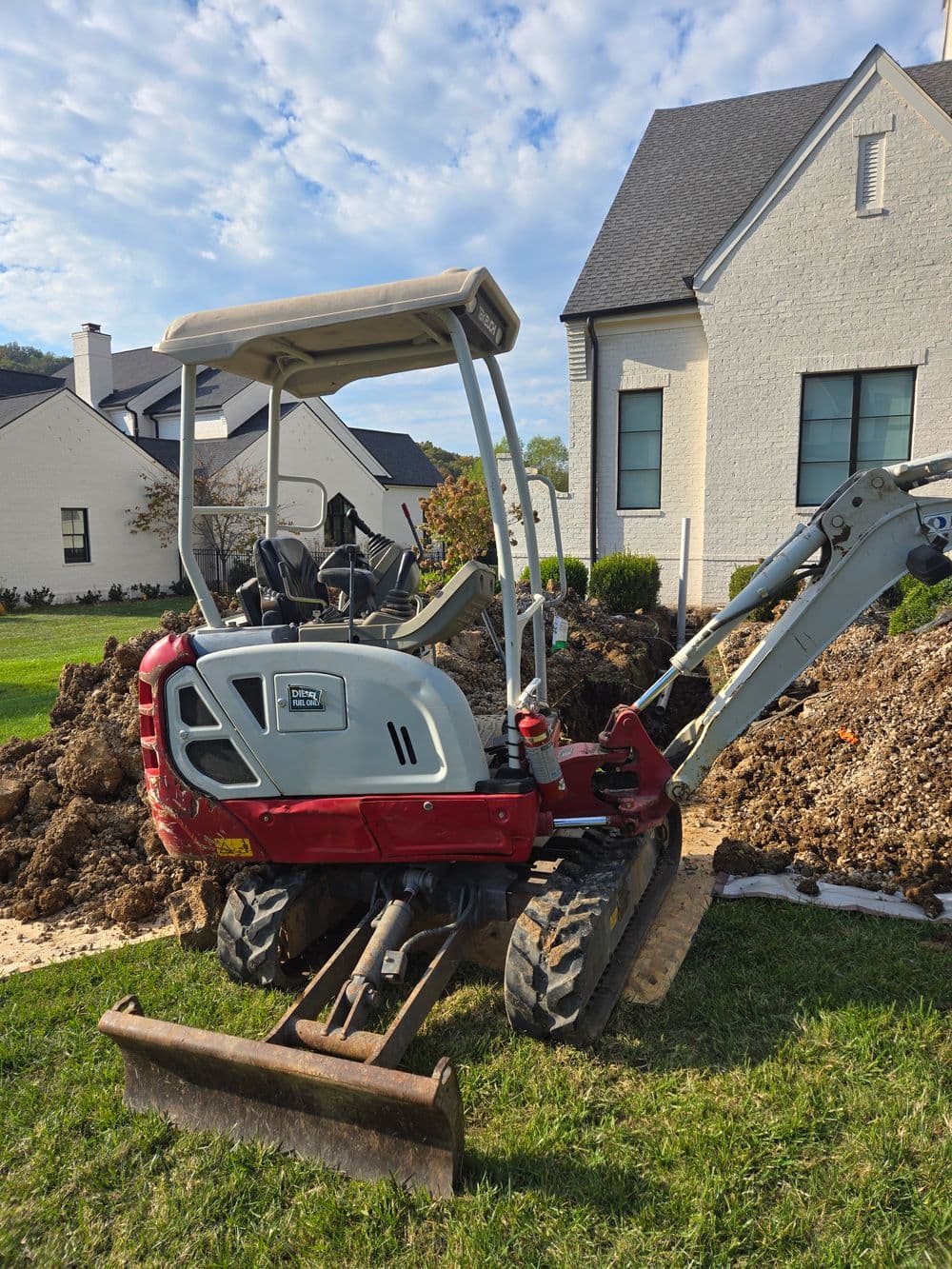 Small excavator parked on a residential lawn, next to a dirt mound and modern home.