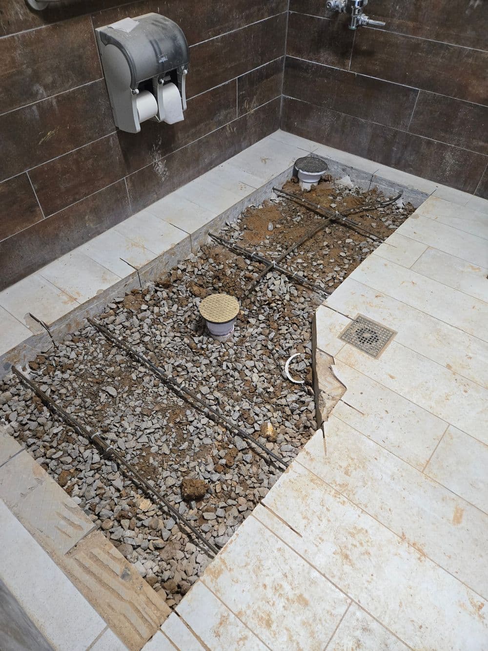 Construction zone in a bathroom showing exposed gravel and plumbing for drainage installation.