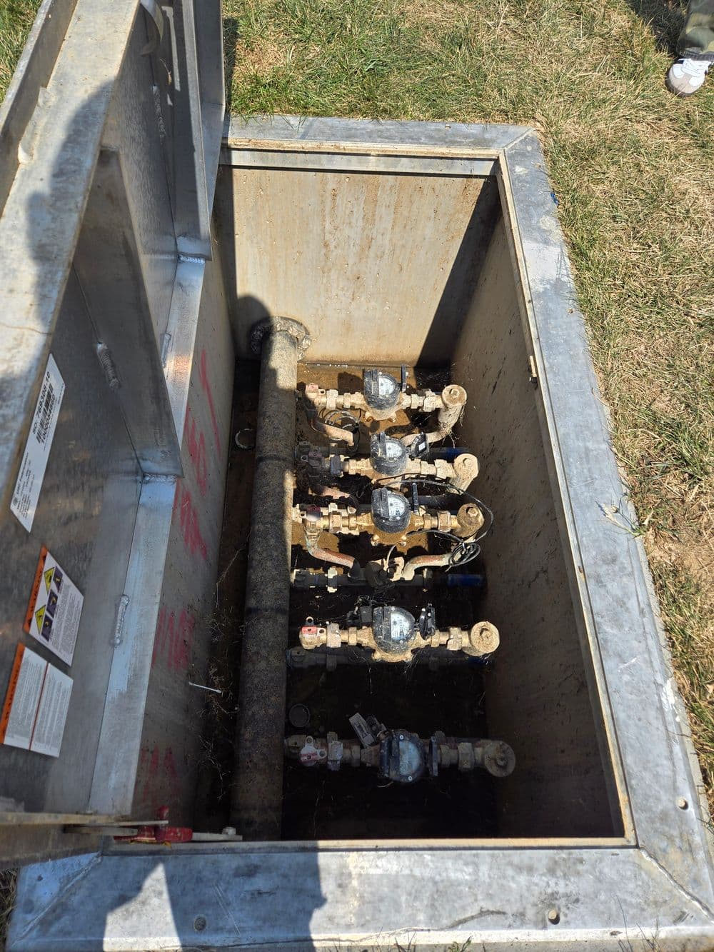 Underground utility valve box with multiple water valves and pipes in grass area.