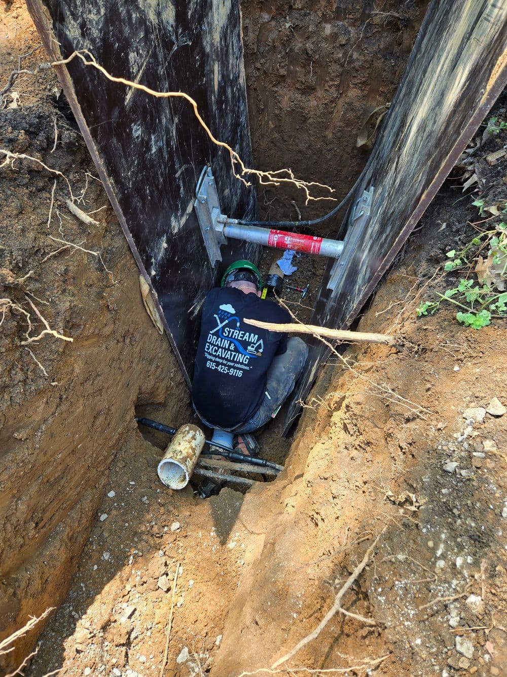 Construction worker inside deep excavation trench installing pipe. Safety gear visible.