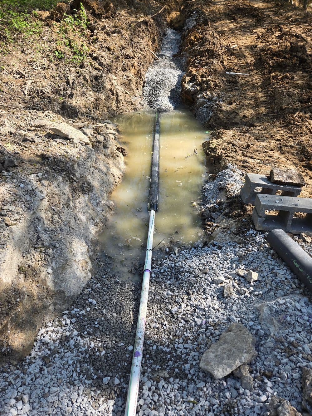 Waterlogged trench with exposed pipe, surrounded by gravel and disturbed earth in a construction site.
