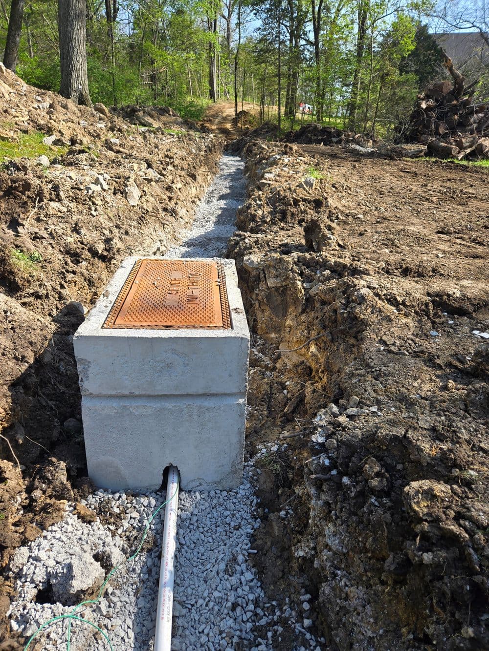 Concrete utility box installed in a dirt trench with gravel, surrounded by trees and clear sky.